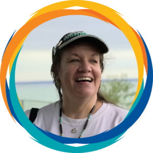 A woman wearing a baseball hat and two necklaces smiles while looking off camera, in front of a blurred body of water.